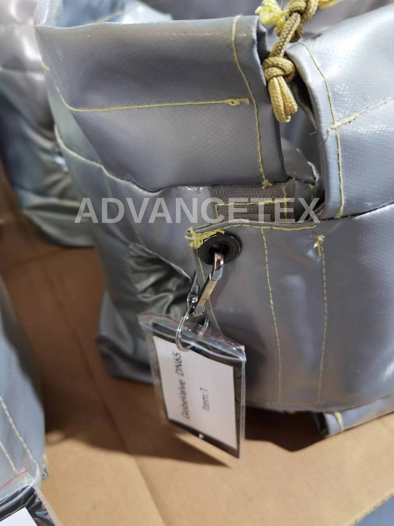 A removable valve jacket from one of our export orders: grey PTFE-coated outer fabric, yellow aramid stitching, hook-and-loop flap, drawcord, eyelet with snap hook, and an item tag reading "GlobeValve DN65 – Item 1".